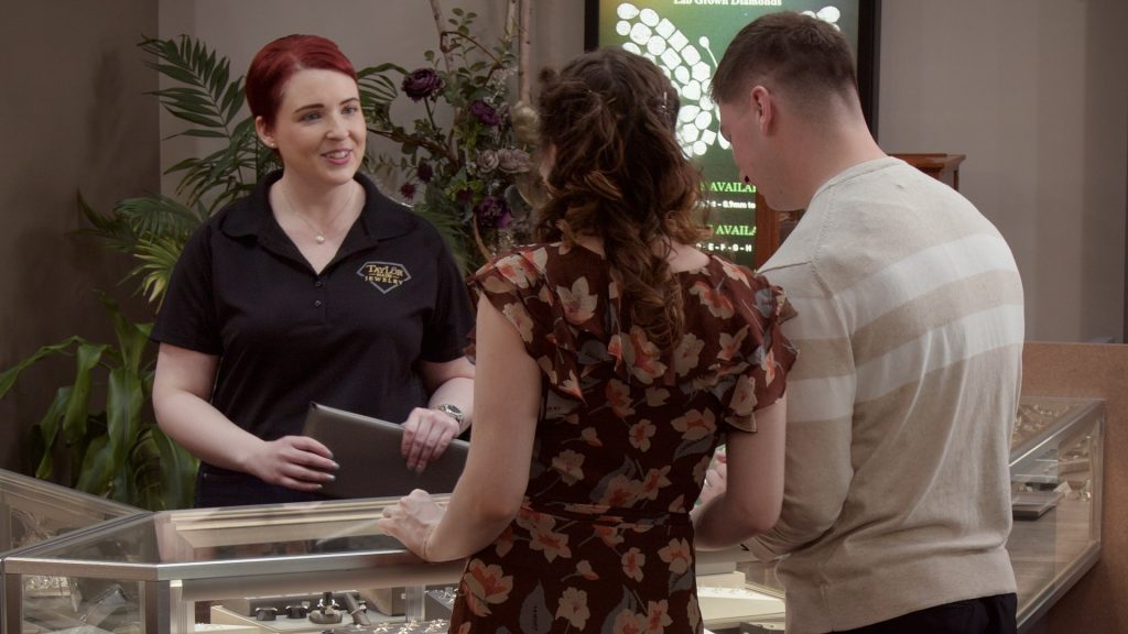 Close-up of a couple shopping for fine jewelry at Taylor Made Jewelry, with a friendly sales associate offering assistance, showcasing engagement rings or wedding bands in the jewelry display case.