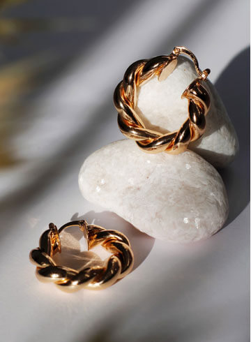 Twisted gold hoop earrings and ring displayed on smooth white stones, showcasing elegant jewelry design and craftsmanship.