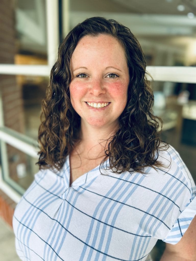 Warm smile woman with curly brown hair wearing a light blue and white striped shirt, capturing a friendly and approachable vibe in a casual setting.