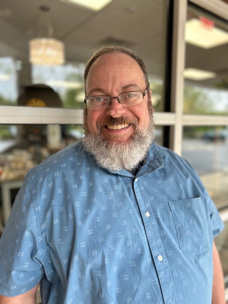 Friendly man with glasses and a gray beard smiling, wearing a light blue patterned shirt, standing inside a jewelry store with blurred background.