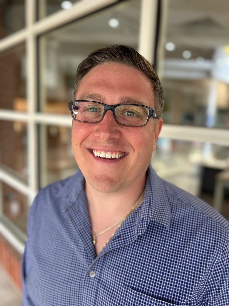 Portrait of a smiling man wearing glasses and a blue checkered shirt, standing inside a jewelry store with shelves filled with jewelry display boxes.